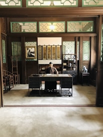 An individual is seated at a wooden desk within a warmly-lit room that features traditional Chinese decor. The desk is elaborately carved, and the individual appears to be engaged in reading or writing. Surrounding the desk are wooden shelves and partitions with intricate lattice designs, filled with various objects such as vases and decorative items. Several framed pictures and scrolls with calligraphy hang on the wall behind the individual.