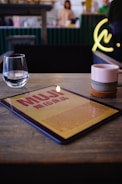A tabletop scene featuring a tablet displaying a page from Muji, with a glass of water and a ceramic mug on the wooden surface. In the background, a blurred interior of what appears to be a cafe can be seen, including counter space and a blurred figure. A neon sign on the wall adds a touch of modern decor.