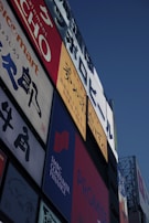 A collection of colorful advertisements and signs on the side of a building, prominently displaying various brands and text in different languages. The signs include Japanese characters and some English text with vibrant red, blue, and yellow backgrounds.