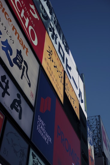 A collection of colorful advertisements and signs on the side of a building, prominently displaying various brands and text in different languages. The signs include Japanese characters and some English text with vibrant red, blue, and yellow backgrounds.