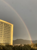 A rainbow arcs across a cloudy sky above a city landscape. To the left, a tall building with numerous windows has the word 'GRAND HYATT' displayed at the top. In the background, mountains are visible, and the foreground includes trees, adding depth to the scene.