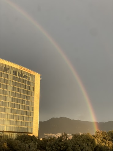 A rainbow arcs across a cloudy sky above a city landscape. To the left, a tall building with numerous windows has the word 'GRAND HYATT' displayed at the top. In the background, mountains are visible, and the foreground includes trees, adding depth to the scene.