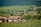 A rural landscape featuring a small village with several wooden houses surrounded by lush greenery. Rolling hills in the background create a serene and peaceful setting. The vegetation includes a field of crops in the foreground and dense forest in the distance.