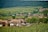 A rural landscape featuring a small village with several wooden houses surrounded by lush greenery. Rolling hills in the background create a serene and peaceful setting. The vegetation includes a field of crops in the foreground and dense forest in the distance.