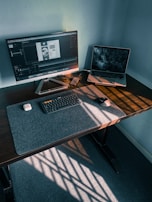 A modern workspace features a large monitor displaying a design application, accompanied by an open laptop showing a dark-themed wallpaper. A keyboard, mouse, wireless earbuds case, and smartphone are neatly arranged on the desk, with sunlight casting striped patterns across the surface.
