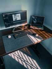 A modern workspace features a large monitor displaying a design application, accompanied by an open laptop showing a dark-themed wallpaper. A keyboard, mouse, wireless earbuds case, and smartphone are neatly arranged on the desk, with sunlight casting striped patterns across the surface.