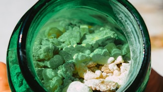 A close-up view of white and light green rocks or granules inside a dark green glass jar. The materials inside the jar appear rough and irregularly shaped, creating a textured appearance.