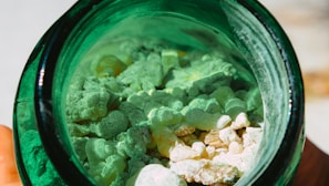 A close-up view of white and light green rocks or granules inside a dark green glass jar. The materials inside the jar appear rough and irregularly shaped, creating a textured appearance.