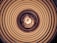A spiraling pattern of concentric circles draws the eye inward towards a glowing light bulb at the center. The circular design creates an optical illusion of depth and movement, enhanced by warm, earthy tones.