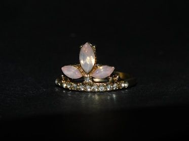 A gold ring adorned with a central arrangement of marquise-shaped moonstones, surrounded by a row of small sparkling diamonds. The setting features a combination of pink and clear stones against a dark background, highlighting the elegance of the jewelry.