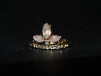A gold ring adorned with a central arrangement of marquise-shaped moonstones, surrounded by a row of small sparkling diamonds. The setting features a combination of pink and clear stones against a dark background, highlighting the elegance of the jewelry.