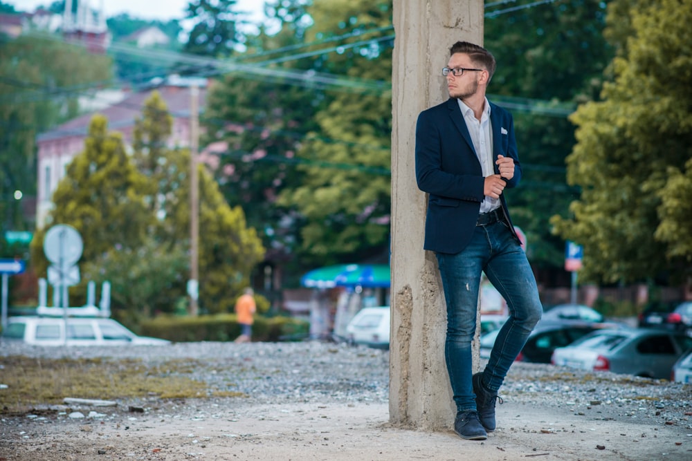 man in black suit jacket and blue denim jeans standing beside brown wooden post during daytime