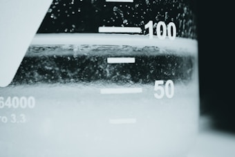 A close-up view of a beaker filled with a clear liquid. The focus is on the measurement markings on the beaker, with numbers like 50 and 100 visible. Bubbles are present within the liquid, and the image has a high contrast between the white of the beaker and the dark liquid.