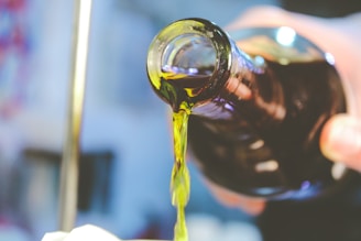 A close-up view of a dark glass bottle as liquid is being poured out from its mouth. The liquid appears to be oil and has a golden hue, suggesting it's olive oil or a similar substance. The background is blurred, focusing attention on the pouring action.