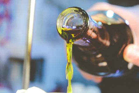 Close-up image of rich golden olive oil pouring into a glass bottle, surrounded by fresh olives and leaves.