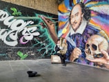A vibrant mural featuring a large portrait with colorful swirls, alongside a hand holding a pen, and another holding a skull. A person dressed in a costume sits nearby playing a tuba, with a hat on the ground and a vintage suitcase beside them. The scene has a dynamic mix of graffiti-style text and artistic design.