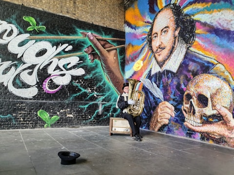 A vibrant mural featuring a large portrait with colorful swirls, alongside a hand holding a pen, and another holding a skull. A person dressed in a costume sits nearby playing a tuba, with a hat on the ground and a vintage suitcase beside them. The scene has a dynamic mix of graffiti-style text and artistic design.