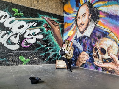 A vibrant mural featuring a large portrait with colorful swirls, alongside a hand holding a pen, and another holding a skull. A person dressed in a costume sits nearby playing a tuba, with a hat on the ground and a vintage suitcase beside them. The scene has a dynamic mix of graffiti-style text and artistic design.