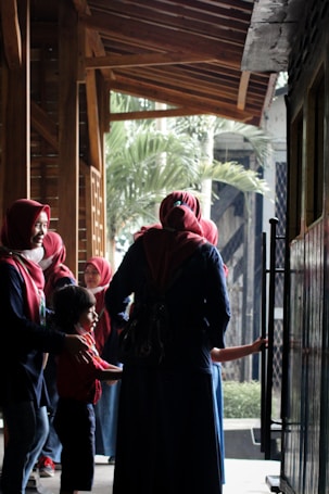 A group of people, predominantly women wearing red hijabs and dark clothing, are standing in a wooden hallway. One of them is interacting with a young child dressed in a red shirt. The background features wooden architecture and green foliage visible through an open area.