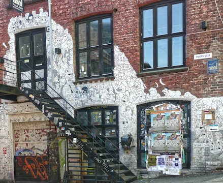 An exterior of a brick building embellished with intricate black and white graffiti art featuring various cartoon-like characters. Windows are present, and a metal staircase leads to a door covered with art. Posters are plastered on a lower door, adding a sense of urban decay and artistic expression.