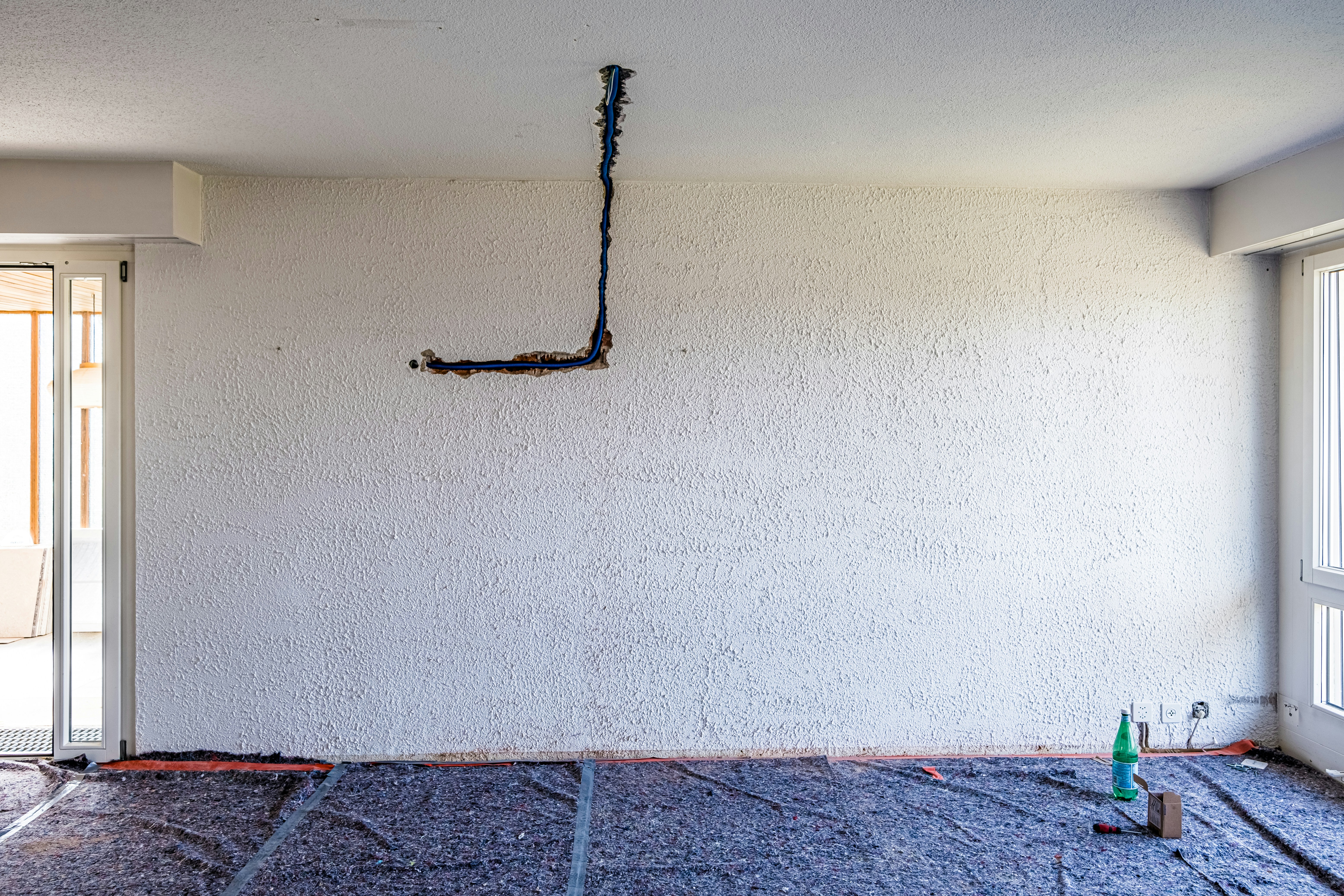 An unfinished interior room with a textured white wall and exposed electrical wiring hanging. The floor appears to be covered with protective material, indicating ongoing renovation work. In the corner, there is a bottle and some tools, suggesting construction or maintenance activities.