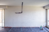 An unfinished interior room with a textured white wall and exposed electrical wiring hanging. The floor appears to be covered with protective material, indicating ongoing renovation work. In the corner, there is a bottle and some tools, suggesting construction or maintenance activities.