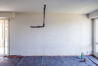 An unfinished interior room with a textured white wall and exposed electrical wiring hanging. The floor appears to be covered with protective material, indicating ongoing renovation work. In the corner, there is a bottle and some tools, suggesting construction or maintenance activities.