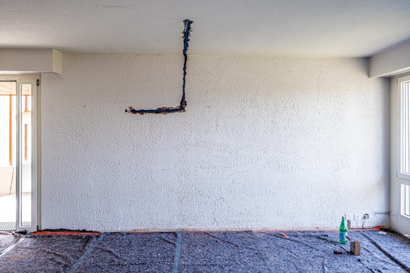 An unfinished interior room with a textured white wall and exposed electrical wiring hanging. The floor appears to be covered with protective material, indicating ongoing renovation work. In the corner, there is a bottle and some tools, suggesting construction or maintenance activities.