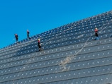 Several workers equipped with safety gear are positioned on the sloped surface of a large structure, possibly engaged in cleaning or maintenance. They are spread out at different levels, with ropes providing support and safety. The sky is clear and vividly blue, adding contrast to the darker surface they are working on.