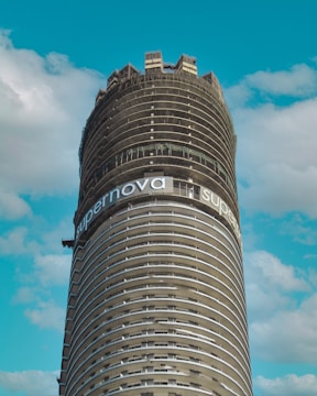A large cylindrical building under construction with numerous horizontal layers. The structure is surrounded by a few sparse clouds against a backdrop of a clear blue sky. At the top portion of the building, there is a banner displaying the word 'supernova'. The unfinished building appears to have scaffolding and other construction materials around the upper section.