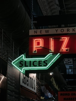 Neon signage with the words 'New York PIZZA Slices' is prominently displayed. The red and green lights contrast sharply against a dark industrial indoor setting. Various menus and additional signage can be seen in the background.