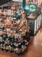 An indoor flower market features a variety of colorful floral arrangements in black plastic buckets. Price tags are visible on the arrangements, and a neon sign reads 'Market Blooms' above a counter displaying additional merchandise. The area is softly lit with hanging lights, creating a warm and inviting atmosphere.