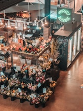 An indoor flower market features a variety of colorful floral arrangements in black plastic buckets. Price tags are visible on the arrangements, and a neon sign reads 'Market Blooms' above a counter displaying additional merchandise. The area is softly lit with hanging lights, creating a warm and inviting atmosphere.