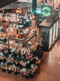 An indoor flower market features a variety of colorful floral arrangements in black plastic buckets. Price tags are visible on the arrangements, and a neon sign reads 'Market Blooms' above a counter displaying additional merchandise. The area is softly lit with hanging lights, creating a warm and inviting atmosphere.