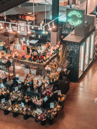An indoor flower market features a variety of colorful floral arrangements in black plastic buckets. Price tags are visible on the arrangements, and a neon sign reads 'Market Blooms' above a counter displaying additional merchandise. The area is softly lit with hanging lights, creating a warm and inviting atmosphere.