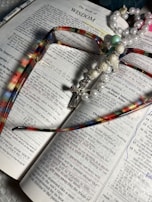 An open book displaying the text of 'The Book of Wisdom' with colorful eyeglasses lying on top. A pearl and bead rosary, featuring a cross, is draped across the book. The pages are marked with underlined sections and handwritten notes in blue ink.