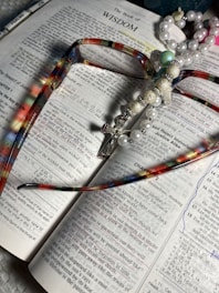 An open book displaying the text of 'The Book of Wisdom' with colorful eyeglasses lying on top. A pearl and bead rosary, featuring a cross, is draped across the book. The pages are marked with underlined sections and handwritten notes in blue ink.