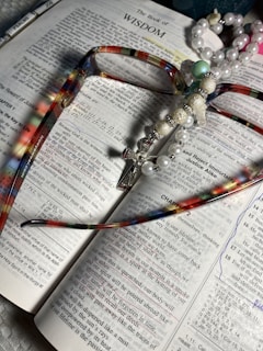 An open book displaying the text of 'The Book of Wisdom' with colorful eyeglasses lying on top. A pearl and bead rosary, featuring a cross, is draped across the book. The pages are marked with underlined sections and handwritten notes in blue ink.