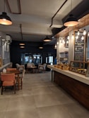 The image captures a cozy cafe interior with dim lighting and a warm atmosphere. Wooden tables and chairs are arranged neatly near a counter displaying a variety of baked goods. Chalkboards with handwritten menus are visible on the walls, and hanging pendant lights provide soft illumination. The overall setting suggests a comfortable space for relaxation and socializing.