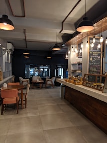 The image captures a cozy cafe interior with dim lighting and a warm atmosphere. Wooden tables and chairs are arranged neatly near a counter displaying a variety of baked goods. Chalkboards with handwritten menus are visible on the walls, and hanging pendant lights provide soft illumination. The overall setting suggests a comfortable space for relaxation and socializing.