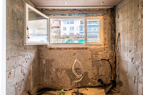 A partially renovated room with bare, unfinished walls showing visible patches of brickwork and mortar. There are exposed pipes and cables on the floor, and a small shovel and a broom leaning against the wall. A large window lets in natural light from outside, showing a view of residential buildings.