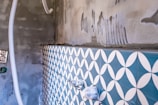 A partially tiled wall with a geometric blue and white pattern, next to an unfinished concrete surface. There are visible plastic pipes and an electrical socket on the left, with some grout and construction markings present.