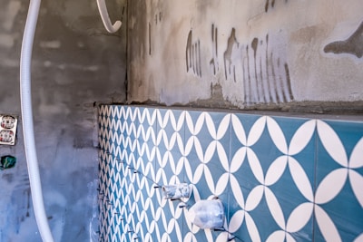 A partially tiled wall with a geometric blue and white pattern, next to an unfinished concrete surface. There are visible plastic pipes and an electrical socket on the left, with some grout and construction markings present.