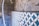 A partially tiled wall with a geometric blue and white pattern, next to an unfinished concrete surface. There are visible plastic pipes and an electrical socket on the left, with some grout and construction markings present.