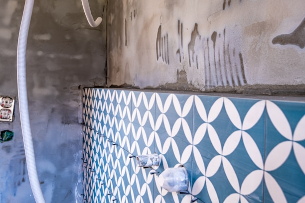 A partially tiled wall with a geometric blue and white pattern, next to an unfinished concrete surface. There are visible plastic pipes and an electrical socket on the left, with some grout and construction markings present.