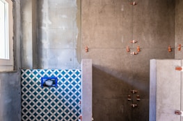 A partially renovated bathroom wall with unfinished surfaces. One section features blue and white geometric tiles, while another section showcases large, grey tiles held in place by orange spacers. There is a window to the left providing natural light.