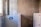A partially renovated bathroom wall with unfinished surfaces. One section features blue and white geometric tiles, while another section showcases large, grey tiles held in place by orange spacers. There is a window to the left providing natural light.