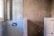 A partially renovated bathroom wall with unfinished surfaces. One section features blue and white geometric tiles, while another section showcases large, grey tiles held in place by orange spacers. There is a window to the left providing natural light.