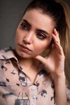 A person gazes thoughtfully to the side with their hand resting on their temple. The individual has expressive eyes, neatly styled hair, and is wearing a patterned shirt with abstract designs and buttons.