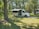 A white recreational vehicle is parked on a grassy area surrounded by a forest of tall trees. The vehicle has its side canopy extended, providing shade. Sunlight filters through the leaves, creating a dappled pattern on the ground.